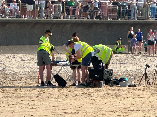 Team members preparing the rocket on a table on the beach, with spectators behind the promenade railings